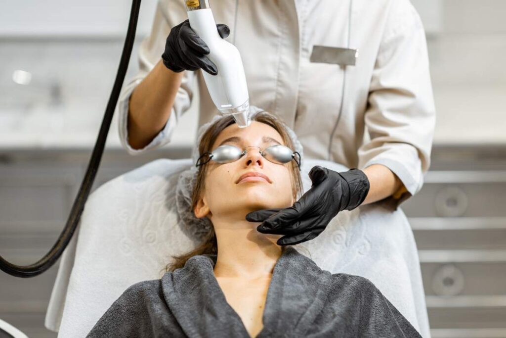A woman with a clean face relaxes in a treatment chair while a clinician gently applies a laser device to her cheek.